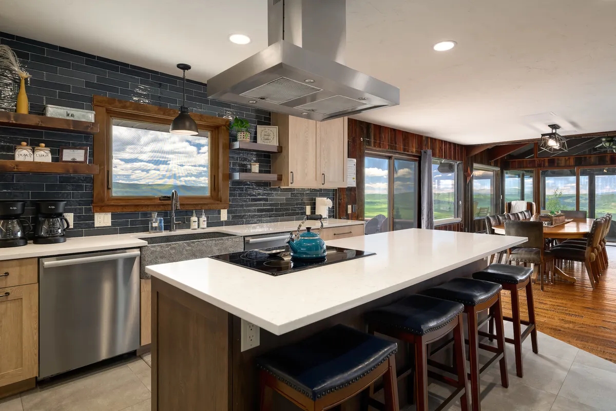 A modern, spacious kitchen featuring a generous island countertop, stainless steel appliances, expansive wooden shelves, and large windows that let in natural light. The foreground highlights a blue kettle, and the open layout is ideal for group cooking and entertaining.