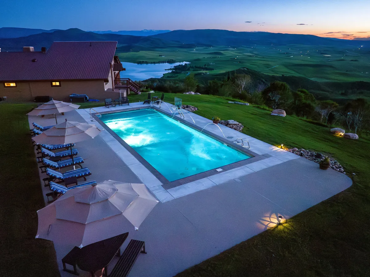 A tranquil outdoor pool area set against a sweeping landscape view, with poolside seating and umbrellas for comfort. The pool is surrounded by greenery and offers a relaxing atmosphere for summer enjoyment, perfect for group gatherings.
