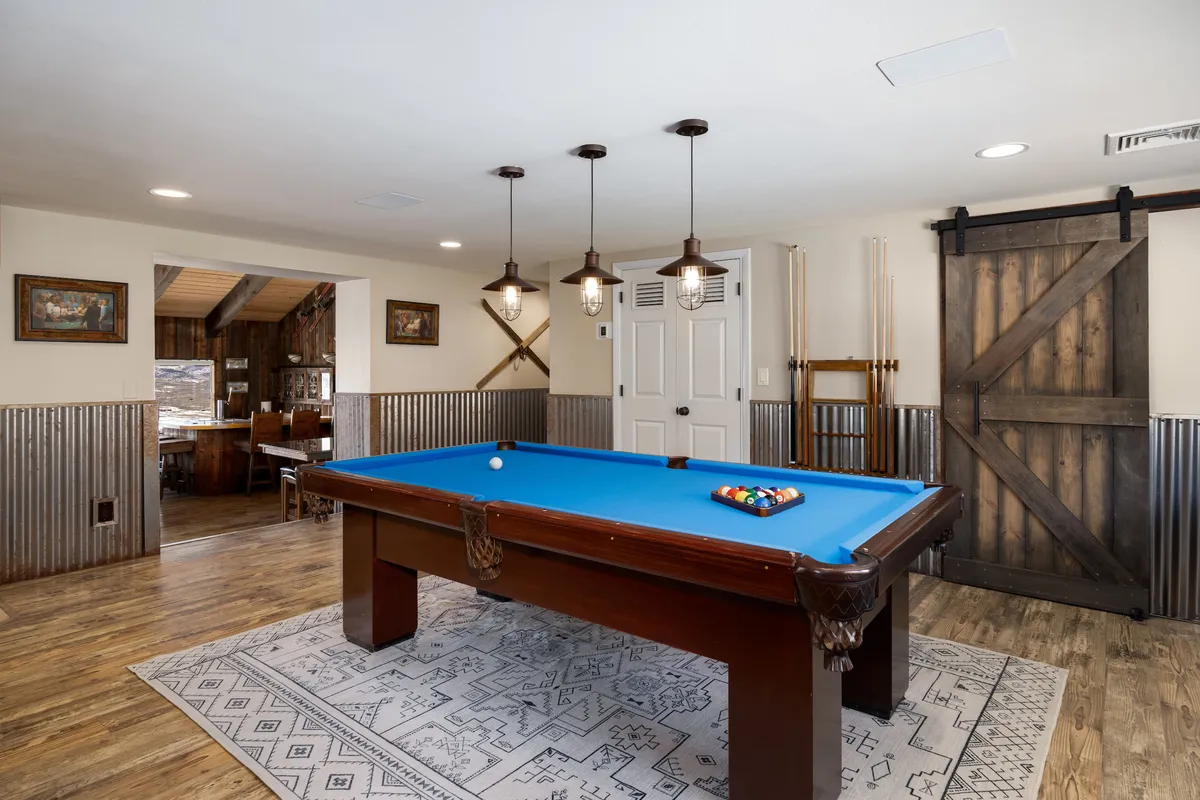 Inside, this image shows a vibrant game room with a pool table, pendant lights, a cozy rug, and a wooden barn door, evoking a playful, communal ambiance. The space is clearly designed for friendly competition and winding down after outdoor activities.