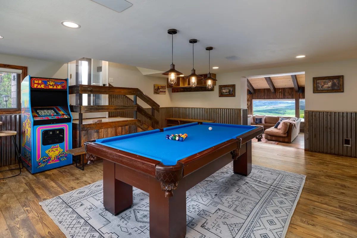 A lively game room with a pool table, an arcade game, cozy seating, and large windows with scenic views. The space is arranged for both relaxation and fun, with natural light flooding in and a mix of entertainment options inviting friendly competition.