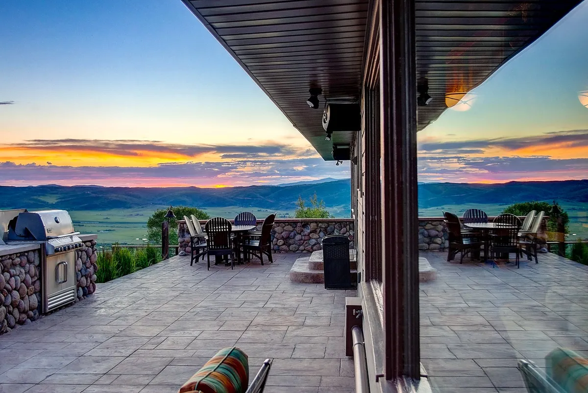 This exterior shot features an outdoor dining area complete with a grill setup, patio flooring, and sweeping mountain vistas. The inviting space is bathed in soft, golden light, suggesting memorable barbecues, celebratory dinners, and relaxed afternoons with friends looking out over the landscape.