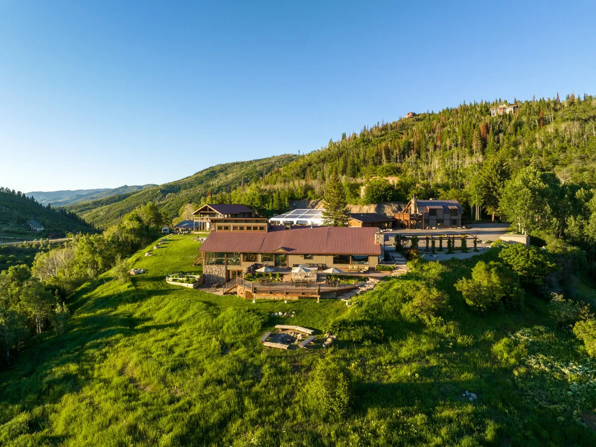 This image captures the main lodge with its expansive deck area set against a backdrop of surrounding greenery and dramatic mountains. The scene suggests ample space for outdoor gatherings, barbecues, and stargazing, with the deck inviting guests to socialize as the sun sets over the mountains.