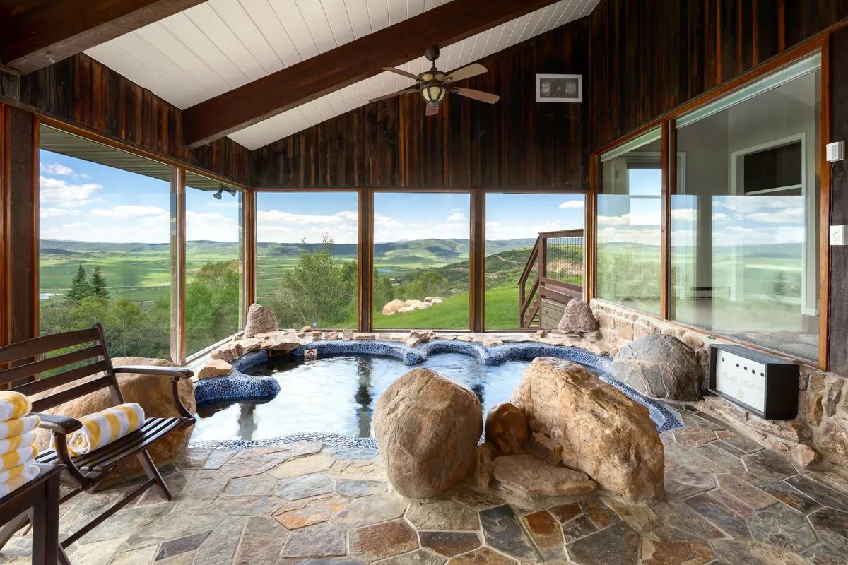 A serene spa pool room featuring natural stone walls, large windows with views of the outdoors, and exposed wooden beams. The tranquil pool is bordered by boulders and the seating area is designed for relaxation and socializing, with sunlight streaming in to create a spa-like ambiance.