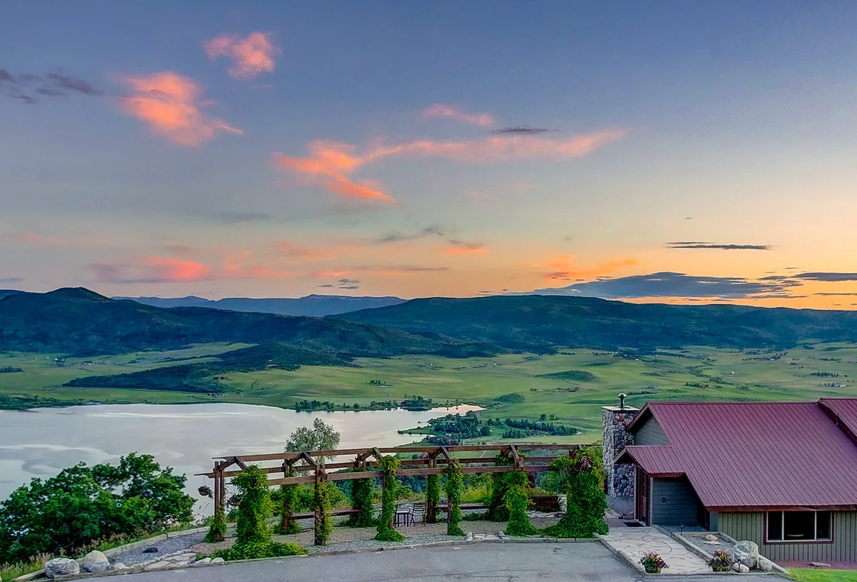 This view captures a serene sunset sky over a tranquil lake, mountains in the distance, and a pergola beside the house. The scene suggests quiet moments of reflection, outdoor dining, and the connection between luxury living and Steamboat’s natural beauty.
