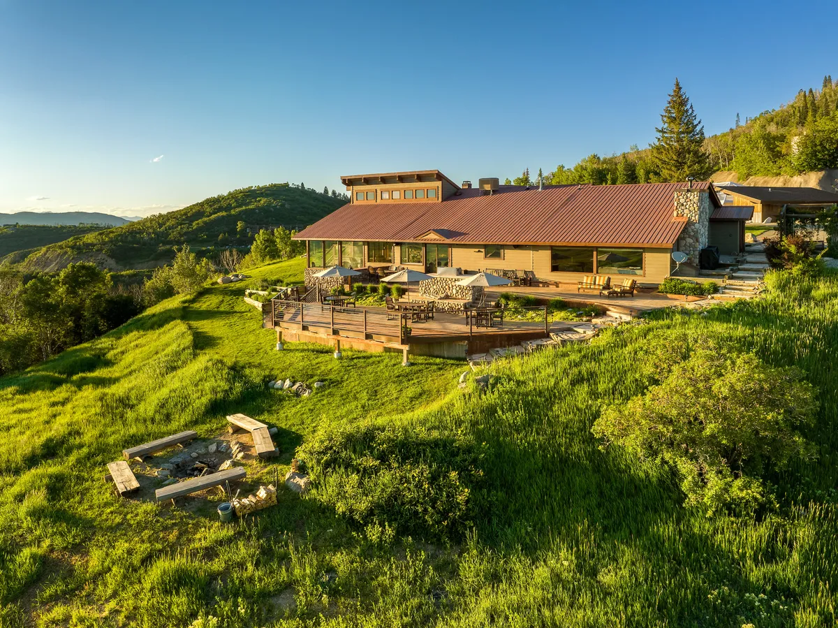 This image showcases the modern house of Bella Vista Lodge with an expansive deck, a cozy fire pit area, and lush greenery all around. The deck offers unobstructed panoramic views of the surrounding mountains and valley, emphasizing the lodge's stunning setting and outdoor luxury.