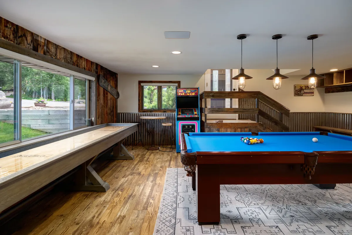 An expansive game space featuring a pool table, shuffleboard table, arcade machine, and large windows. The room is bright and designed for group activities, with distinct gaming features and relaxed seating for guests to socialize and play.