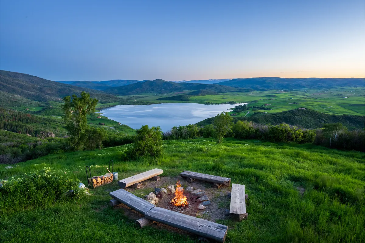 The image shows an inviting exterior setting with a fire pit surrounded by chairs, a scenic lake, expansive mountain views, and lush greenery. The atmosphere is relaxed and peaceful, perfect for group gatherings or unwinding after team-building activities.