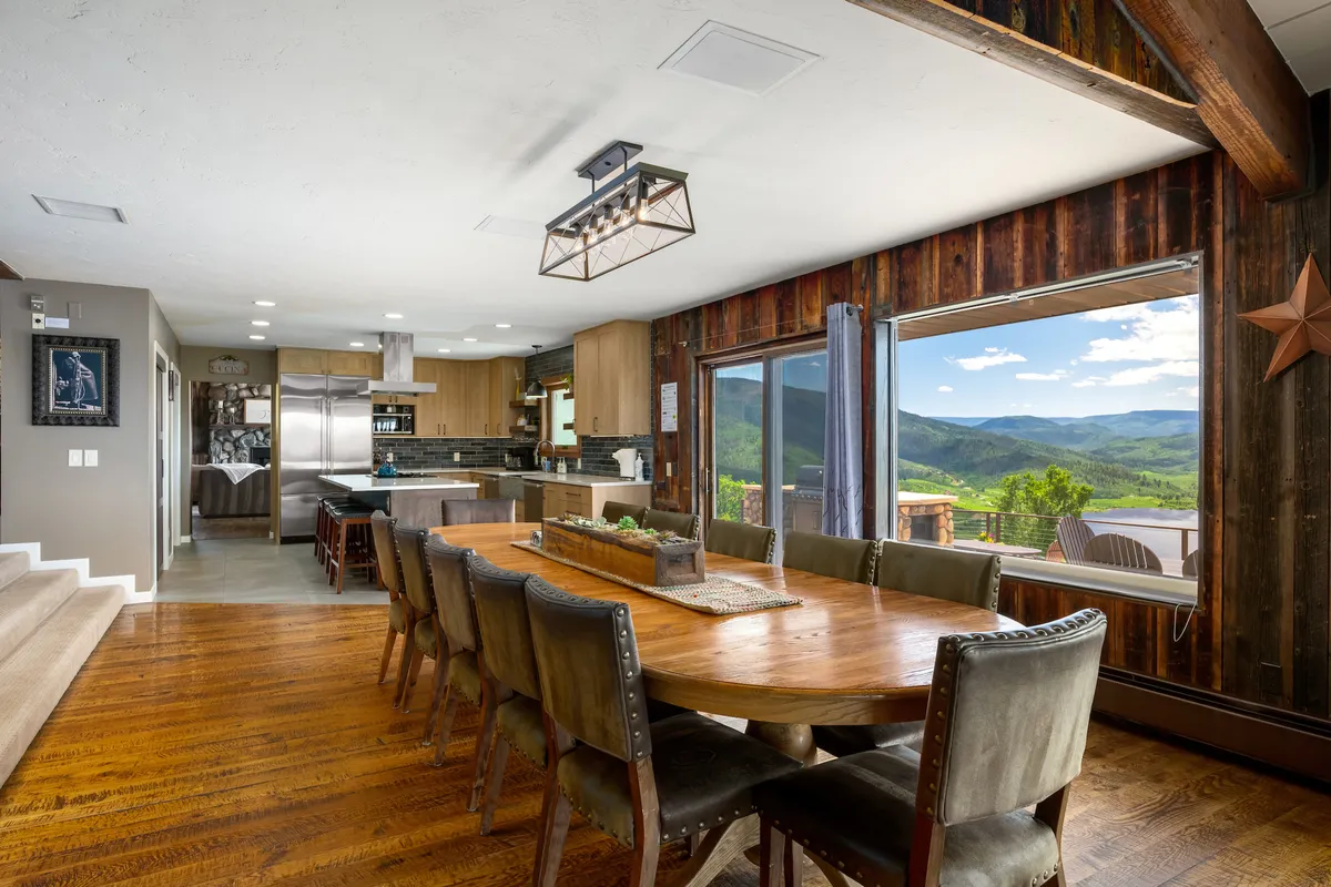 A large dining table set next to panoramic windows, offering a sweeping view of the outdoors. The modern kitchen appliances are visible in the background, connecting the dining and kitchen spaces within a single, inviting open-concept area.