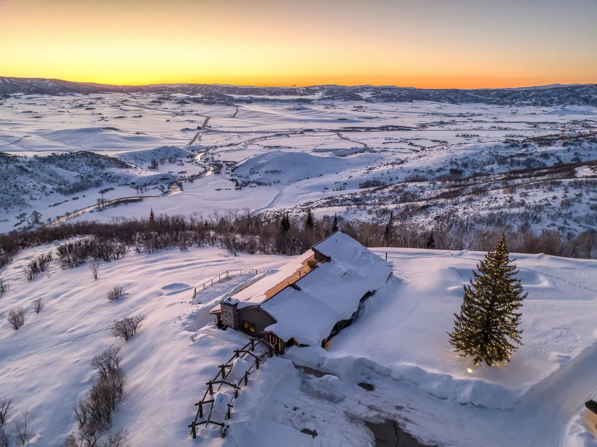 A serene winter exterior of Bella Vista Lodge with a snow-covered landscape, the house nestled among pine trees, and a sunset sky in the background. The scene evokes the magic of wintertime, with opportunities for skiing, snowshoeing, and enjoying cozy evenings after a day outdoors.