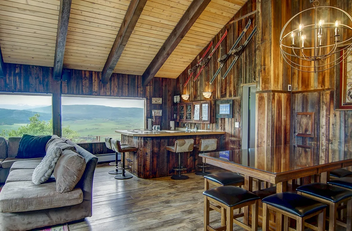 A rustic, inviting bar area with wooden accents, high-top bar seating, ski-themed décor, and large windows offering sweeping mountain views. The space blends relaxation with the excitement of après-ski or evening gatherings, and emphasizes scenic vistas.