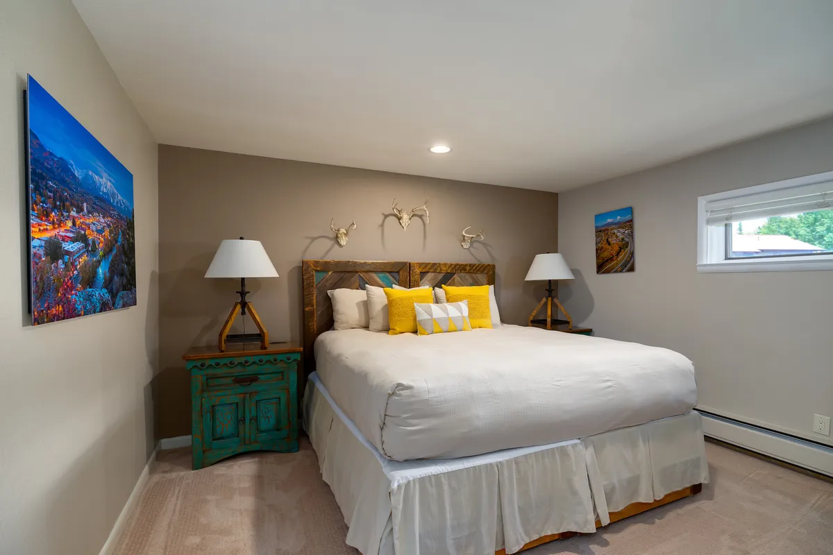 A cozy bedroom with a comfortable bed topped with colorful pillows, decorative lamps, and artwork on the walls. Soft natural light filters into the room, creating a welcoming, restful vibe.