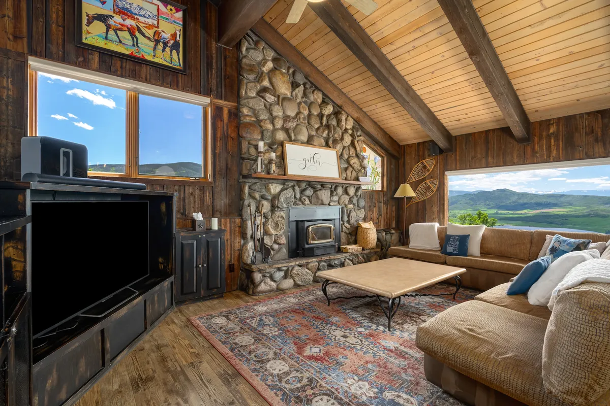 A spacious great room featuring a stately stone fireplace, a large sectional sofa, vaulted ceilings with wooden beams, expansive windows showcasing breathtaking mountain views, and colorful artwork. The room exudes warmth and is designed for groups to gather comfortably around the fire or gaze at the scenery.