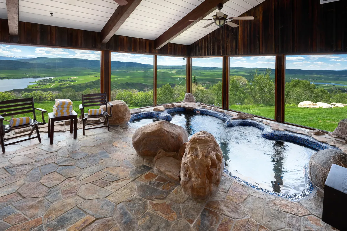 The image showcases an inviting indoor pool surrounded by natural rock formations and elegant lounge chairs. Expansive floor-to-ceiling windows frame breathtaking sunset views over the snow-capped mountains. The wooden ceiling and warm lighting create a cozy, serene atmosphere, perfect for relaxation and social gatherings.